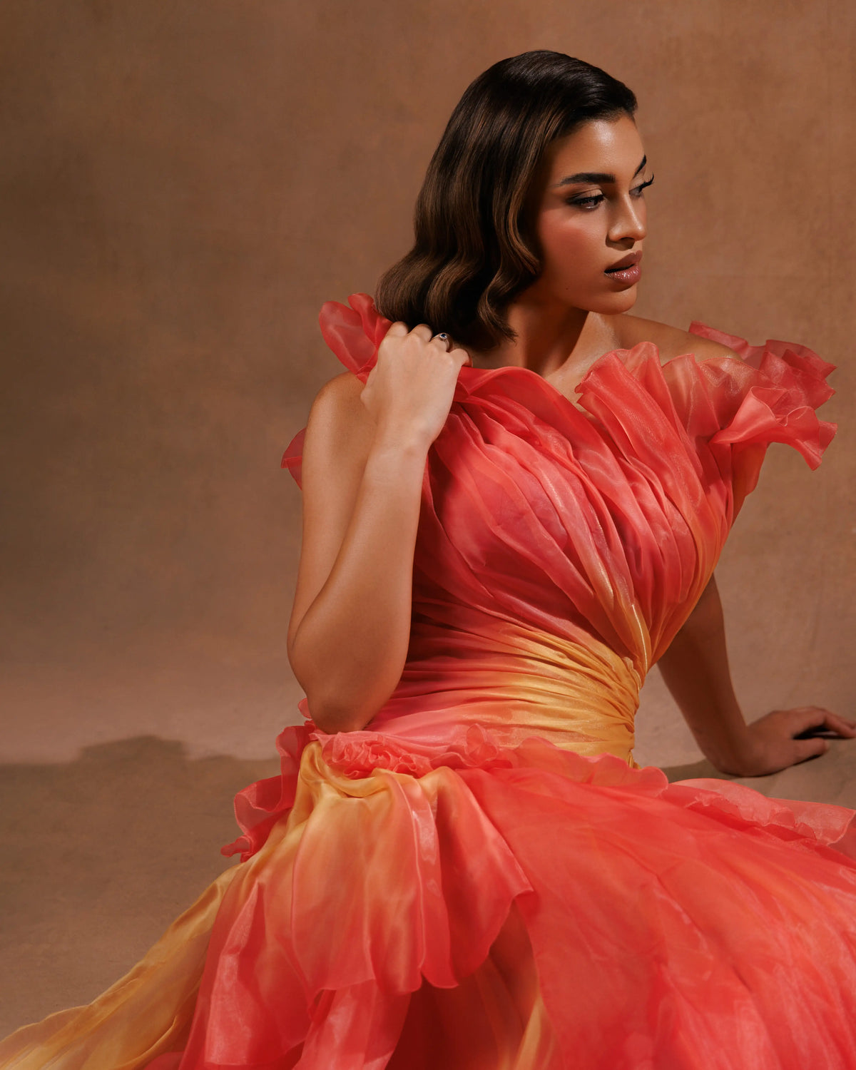 A woman wearing a voluminous ruffled gown in a warm coral-red and yellow gradient, posed against a textured brown studio backdrop.